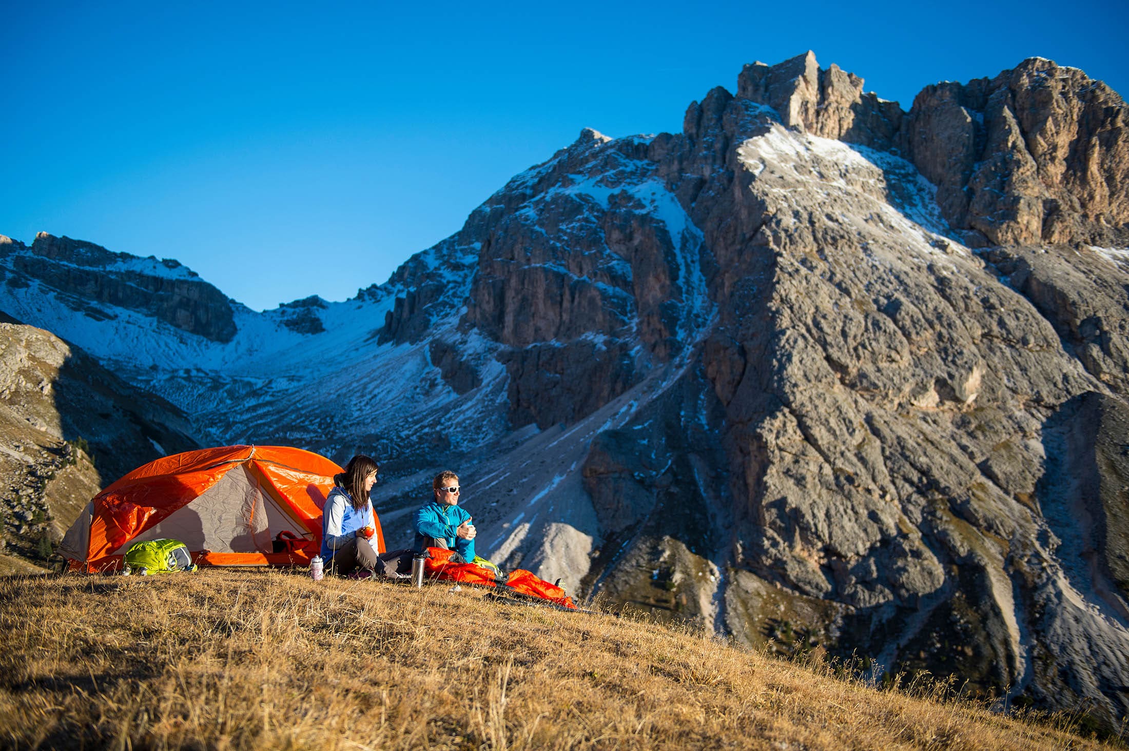 a couple sits outside their tent amid snow-covered mountains